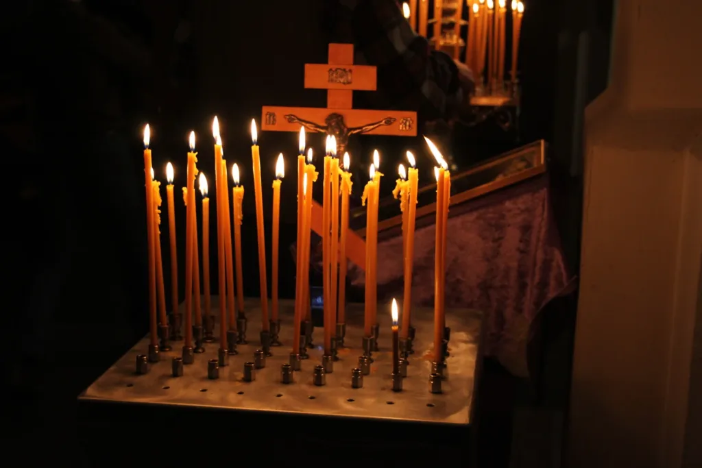 Thin beeswax candles burning on a candle stand during the Divine Liturgy, a wooden cross on the stand behind them.