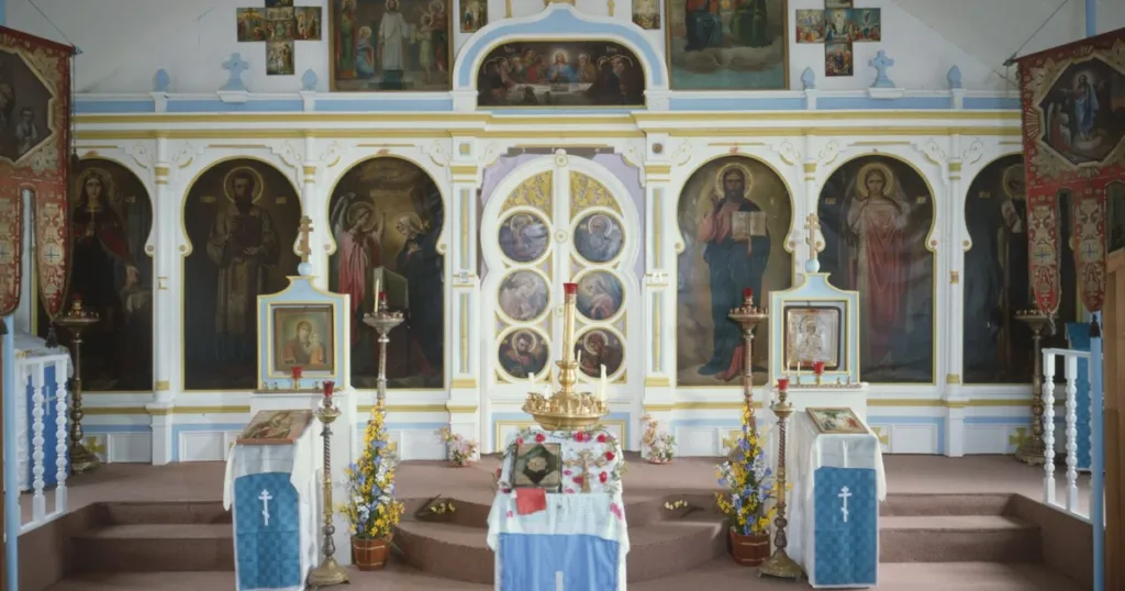 The nave of a small wooden Orthodox church, looking east to a white and blue iconostasis with large icons of Christ, the Theotokos and the saints.