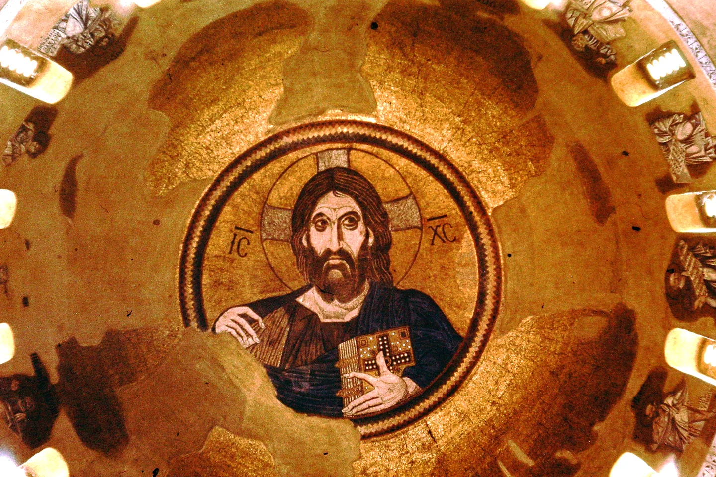 A mosaic of Christ Pantocrator, holding the Gospel book, in the center of a golden church dome.