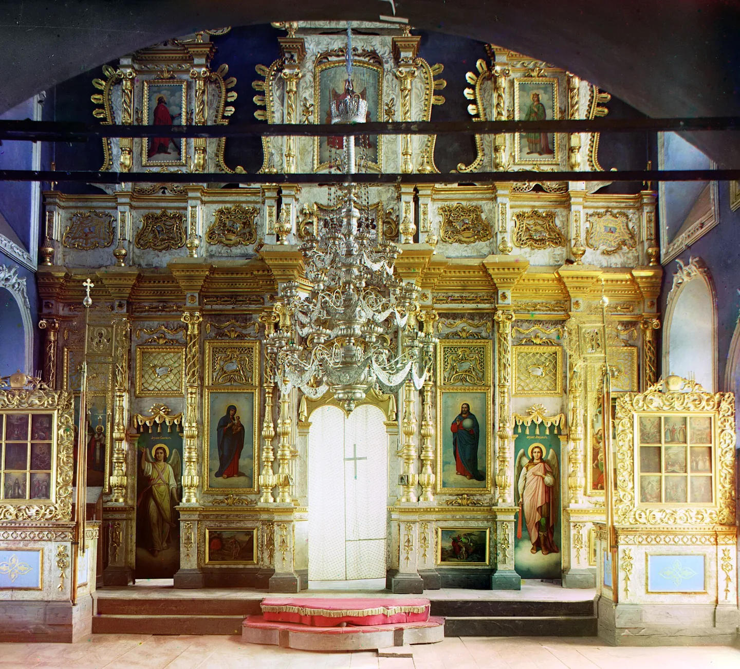 A tall gilded iconostasis with tiers of icons and the Royal Doors in the center, a chandelier hanging in front of it.